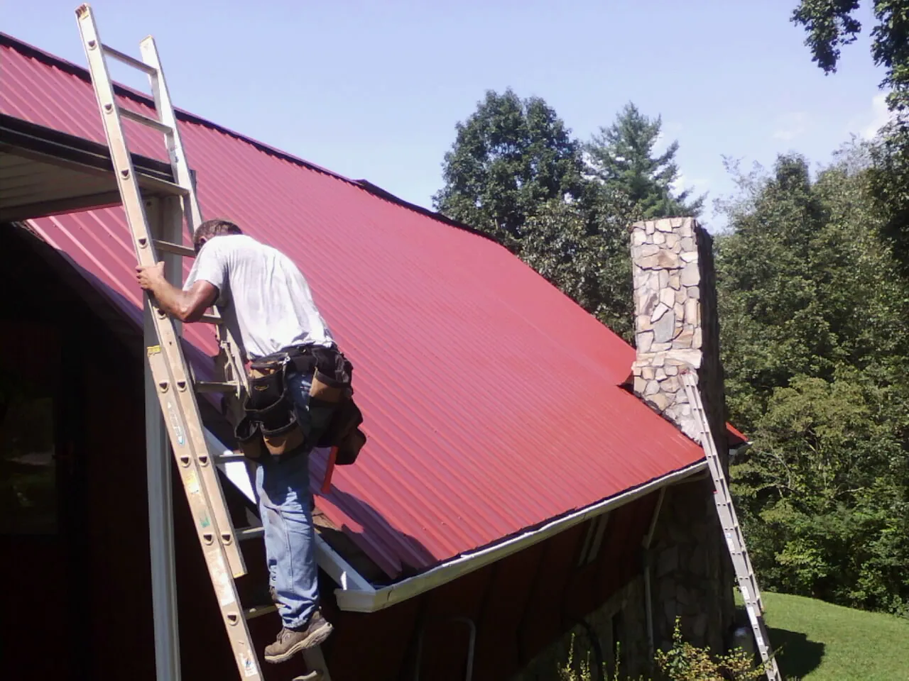 Roofer on a ladder beside a red metal roof and stone chimney — Soffit & Fascia Repair by Roofing Brothers in Ada